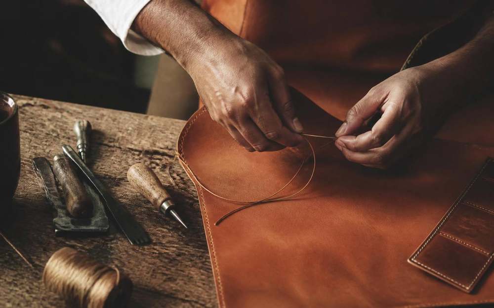Indian artisan hand-stitching premium cognac leather at a wooden workbench
